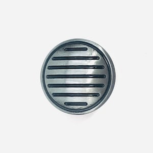 May include: A circular metal knob with a brushed silver finish, featuring a series of horizontal black recessed lines across the face. The knob has a smooth, rounded outer edge and a flat, disc-like profile, presenting a clean and industrial design.