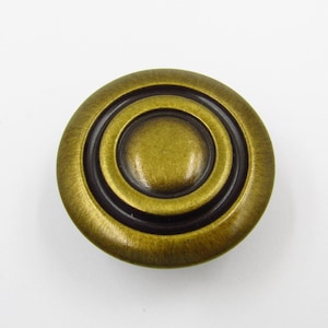 Może przedstawiać: Okrągła gałka do szafki w kolorze antycznego mosiądzu z koncentrycznymi okręgami. Gałka ma podniesiony środek i ciemny, zagłębiony pierścień między zewnętrznym i wewnętrznym okręgiem. Wykończenie ma lekko teksturowany wygląd. Idealna do renowacji mebli.