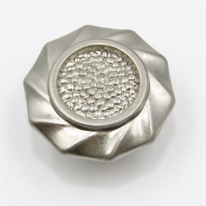May include: A silver-toned cabinet knob with a textured, circular design. The knob has a starburst pattern around the edge.