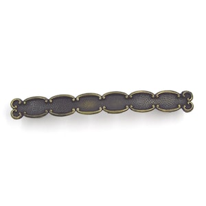 May include: A decorative, antique-style cabinet pull handle. The handle is dark bronze with a series of oval shapes connected in a row. The edges of the ovals are outlined in a lighter gold color. The handle has decorative flourishes on each end.