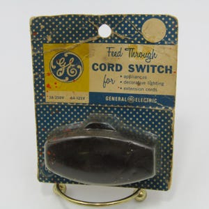 Puede incluir: Interruptor de cable General Electric vintage en su embalaje original. El embalaje es azul con lunares blancos y presenta el logotipo de GE. El texto dice "Feed Through Cord Switch" y enumera aplicaciones. El interruptor es marrón oscuro.