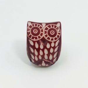 May include: A deep red, translucent owl-shaped figurine featuring carved, white-filled circular eye patterns and a series of white-filled, teardrop-shaped feather details across the body, creating a high-contrast, textured appearance.