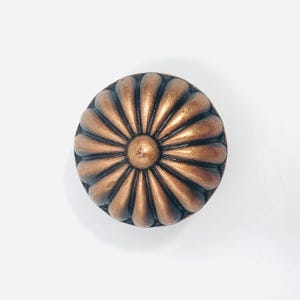 May include: A round cabinet knob with a metallic copper finish, featuring a raised, radial petal pattern that radiates from a central circular point. The design consists of twelve symmetrical, elongated, curved segments.