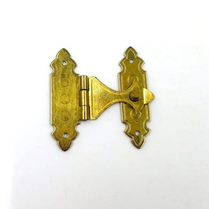 May include: A brass-coloured metal hinge featuring two elongated plates with decorative scalloped edges and circular cutouts. The left plate is rectangular with three circular indentations, while the right plate has a pointed, arrow-like extension.