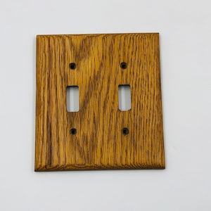 May include: A rectangular light brown wooden double light switch wall plate featuring a prominent natural wood grain pattern. The plate includes two vertical rectangular cutouts for toggle switches and four small circular screw holes for mounting.