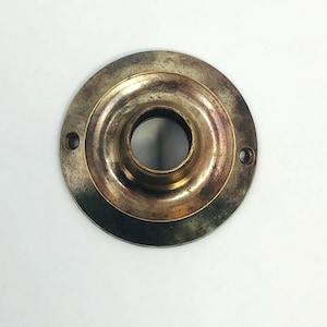 Op de afbeelding: Een vintage, cirkelvormig metalen object met een centraal gat en twee kleinere gaten. Het metaal heeft een aangetaste, verouderde uitstraling met goud- en bruintinten, wat suggereert dat het een antiek of gerecycled item is.