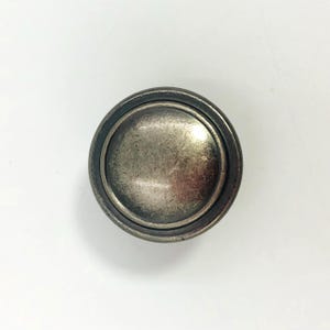 May include: A circular cabinet knob with a distressed, antique silver finish. The knob features a smooth, slightly domed center surrounded by a raised, darker metallic rim, creating a layered, dimensional appearance with visible texture.