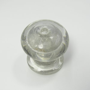 Puede incluir: Un pomo de vidrio transparente con una parte superior redondeada y una base cilíndrica. El pomo tiene un pequeño agujero en la parte superior y parece ser vintage. El vidrio tiene un aspecto ligeramente turbio.