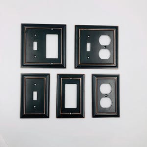 May include: A collection of five rectangular wall plates in matte black with copper-toned trim. The set includes two double-gang plates and three single-gang plates featuring various configurations for light switches and electrical outlets.