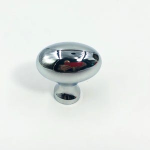 May include: A polished chrome cabinet knob with a smooth, oval-shaped head and a tapered cylindrical base. The metallic surface is highly reflective, showing a distorted mirror image of the surroundings on its rounded, convex top surface.