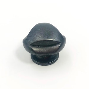 May include: A dark, metallic cabinet knob with a smooth, rounded dome top and a horizontal, recessed groove detail. The hardware features a circular base and a weathered, gunmetal-toned finish with subtle variations in color across the surface.