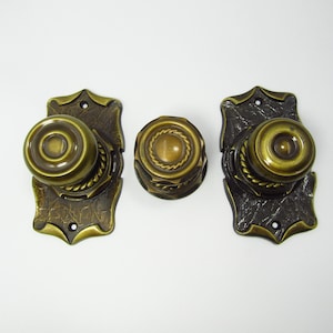 May include: Three antique brass door knobs and plates. The knobs are round with a rope-like design around the center. The plates are ornate with a leaf-like pattern. The finish is a dark brass color.