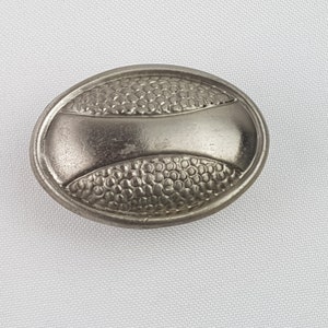 Może przedstawiać: Srebrny, owalny guzik z fakturowaną powierzchnią i wypukłym wzorem. Guzik ma gładki, lekko wypukły środkowy element.