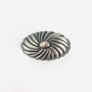 May include: An oval-shaped metal cabinet knob with a brushed silver finish. The surface features a series of raised, radiating ridges that spiral outward from a smooth, rounded center point, creating a textured, fan-like pattern across the piece.