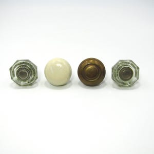 May include: Four antique door knobs in a row. Two are clear glass with an octagonal shape, one is a round, off-white knob, and the other is a round, brass knob. All are set against a white background.