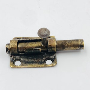 May include: A vintage brass sliding bolt latch with a round knob. The latch has a rectangular base with two screw holes and a cylindrical bolt. The metal has a tarnished, aged appearance.