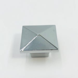 May include: A square cabinet knob with a polished chrome finish. The knob features a pyramid-shaped top with four beveled triangular sides that meet at a central point, attached to a square base that tapers slightly toward the bottom mounting point.