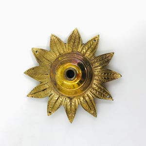 Könnte beinhalten: Ein Vintage-Dekostück in Form einer Sonnenblume aus Messing. Das Stück hat ein zentrales kreisförmiges Element mit einem dunklen Loch, umgeben von strahlenförmigen Blütenblättern. Das Messing hat einen goldenen Farbton mit dunkleren Bereichen.