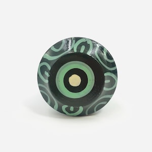May include: A round cabinet knob featuring a dark green base with a light green circular center and a small cream-colored dot in the middle. The outer edge of the knob is decorated with a repeating pattern of light green curved lines.
