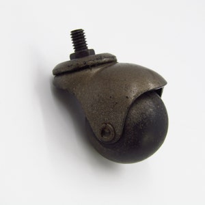 May include: A single antique-style caster wheel with a dark, weathered finish. The wheel has a rounded, black rubber ball and a threaded bolt for attachment. The metal components show signs of age and wear, suggesting a vintage or repurposed item.