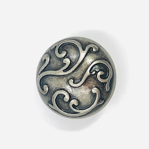 May include: A round, metallic cabinet knob with a dark, antiqued silver finish. The surface features a raised, swirling scrollwork pattern that creates a textured, dimensional appearance across the circular face of the hardware piece.