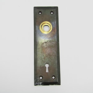 Könnte beinhalten: Rechteckige antike Türplatte mit Schlüsselloch und kreisförmiger Öffnung. Die Platte ist dunkel mit einem angelaufenen Finish und hat vier Schraubenlöcher. Die kreisförmige Öffnung hat einen goldfarbenen Ring.