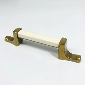 May include: A cabinet pull with a rectangular white center and brass-colored ends. The brass ends have a curved, hook-like design. The pull is designed for easy gripping and opening of drawers or cabinets.