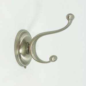 May include: A brushed nickel double hook wall mount. The hook has a decorative circular base and two curved arms, each ending in a small ball. The fixture is designed for hanging items and has a classic, elegant style.
