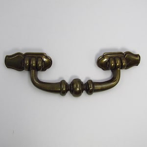 May include: A brass cabinet pull with a decorative design. The pull has a curved shape with two ornate ends and a center knob.