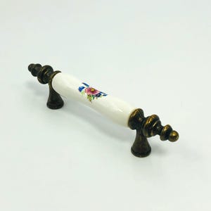 Puede incluir: Un tirador decorativo para armario con un mango de cerámica blanca con un diseño floral en rosa, azul y verde. El tirador tiene detalles metálicos de color bronce y un diseño clásico y ornamentado.