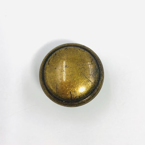 May include: A round, antique brass knob with a slightly domed surface. The knob has a dark, aged patina with visible scratches, suggesting age and use. The edges are rounded, and the overall design is simple and functional.