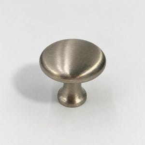 Puede incluir: Un pomo redondo para armario con acabado en níquel cepillado. El herraje presenta una superficie superior circular lisa y ligeramente abombada que se transforma en una base cilíndrica cónica, ofreciendo un diseño limpio y funcional.
