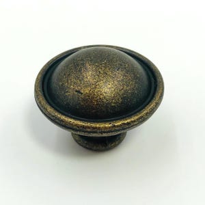 May include: A bronze-colored cabinet knob with a rounded, dome-shaped top. The knob has a textured finish and a dark ring around the base of the dome. The base of the knob is a smaller, rounded shape.