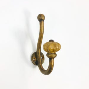 May include: A wall-mounted coat hook featuring a curved brass-toned metal frame with two prongs. The lower, shorter prong is topped with a round, fluted, beige ceramic knob. The base plate includes two screw holes for secure attachment to a wall surface.