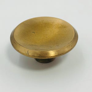 Puede incluir: Un pomo circular para muebles con una superficie superior cóncava y lisa, con un acabado dorado metálico. El pomo presenta un borde exterior ligeramente elevado y redondeado, junto con un punto de fijación central en la base.