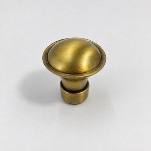 May include: A round cabinet knob with a brushed antique brass finish. The knob features a smooth, domed top surface that transitions into a slightly tapered cylindrical base, providing a functional grip for opening drawers or cabinet doors.