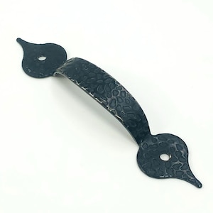 Può includere: Una maniglia per porta in metallo nero martellato con un'impugnatura strutturata e estremità decorative a forma di goccia. L'hardware presenta una finitura scura e un design rustico, adatto per armadi o porte.