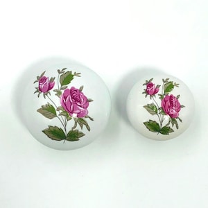 May include: Two white ceramic cabinet knobs featuring a pink rose design with green leaves and stems. The knobs are of different sizes, one larger than the other. The design is a classic floral pattern.