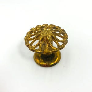May include: A vintage brass cabinet knob with an ornate, openwork floral design. The knob has a round base and a decorative, flower-like top. The brass has a warm, aged patina, suggesting it is an antique or vintage piece.