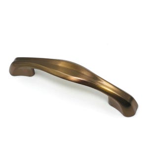 May include: A curved cabinet handle with a smooth, metallic bronze finish. The handle features a tapered, arched design that is wider in the center and narrows toward the two mounting points on each end, showcasing a polished, reflective surface.