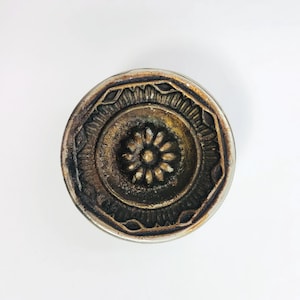Könnte beinhalten: Ein rundes Metallobjekt im antiken Stil mit einem zentralen Blumendesign. Das Objekt hat einen silberfarbenen Rand und eine verwitterte, bronzefarbene Mitte mit aufwendigen Details. Das Design zeigt eine zentrale Blume, umgeben von konzentrischen Kreisen und geometrischen Mustern.