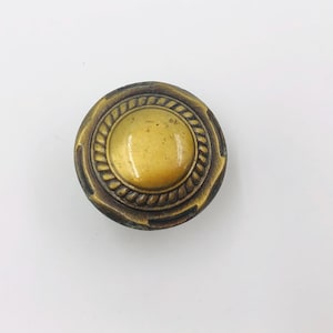 Könnte beinhalten: Ein runder Schrankknopf aus Messing mit einer glatten, gewölbten Mitte, die von einem strukturierten Rand im Seil-Design umgeben ist. Das Metall hat ein dunkles, gealtertes Finish mit sichtbaren Glanzlichtern an den Kanten.
