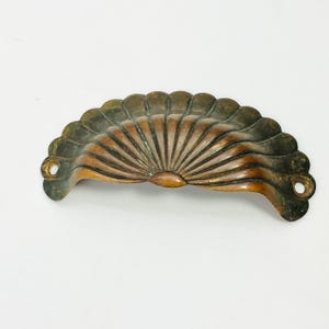 May include: A vintage, fan-shaped drawer pull with a copper finish. The pull has a scalloped edge and a central, radiating design. Two holes are on each end for mounting. The hardware is likely made of metal.