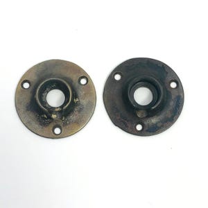 以下が含まれることがあります： 2つのアンティークの円形金属製ドア金具。各部品には、中央に円形の開口部があり、周囲に4つの小さな穴があります。金属は、茶色とブロンズの色合いで、変色した、古くなった外観をしています。