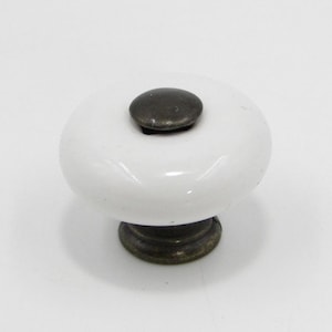 May include: White ceramic cabinet knob with a dark metal center and base. The knob has a rounded, smooth shape, and the metal components have a slightly aged appearance. A decorative hardware element for furniture.