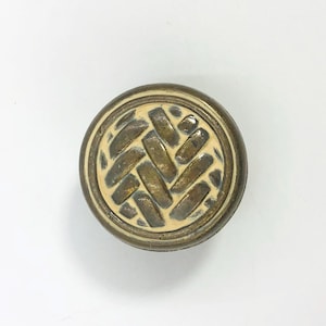 May include: A circular cabinet knob featuring a metallic brass-toned outer rim and a central inlay with a raised, dark bronze-coloured herringbone pattern set against a smooth, cream-coloured background, creating a textured, multi-dimensional appearance.