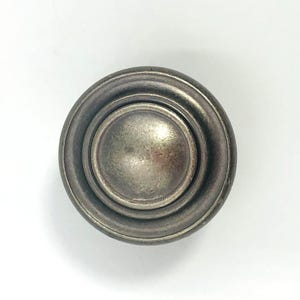 May include: A circular cabinet knob with a weathered, dark metallic finish. The hardware features a raised, smooth central dome surrounded by a concentric ring design, creating a layered, stepped appearance with a muted, antique-style texture.