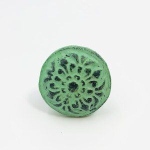 May include: A round cabinet knob featuring a mint green finish with dark, distressed accents. The surface displays a raised, symmetrical floral pattern with a central circular element and radiating petals, creating a textured, weathered appearance.