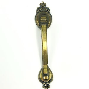 May include: Antique brass-toned drawer pull with ornate detailing. The pull features a decorative top and bottom with a central handle. The hardware is designed for furniture and cabinets, adding a vintage touch to any piece.