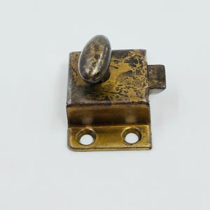 May include: An antique metal latch with a tarnished brass finish. The latch has an oval handle and a rectangular body with two screw holes. The metal shows signs of age and wear, adding to its vintage appeal.